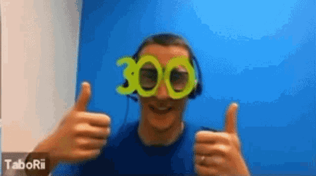 a man wearing headphones and glasses with the number 300 on them gives two thumbs up