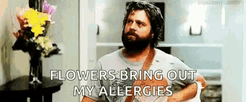 a man with a beard is standing in front of a vase of flowers and saying flowers bring out my allergies .