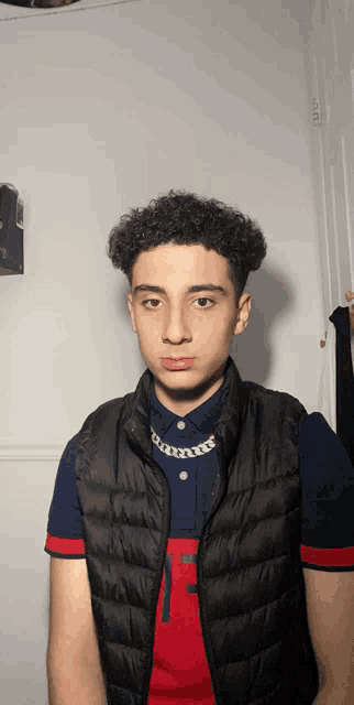 a young man with curly hair wears a black vest and a red shirt with the letter f on it
