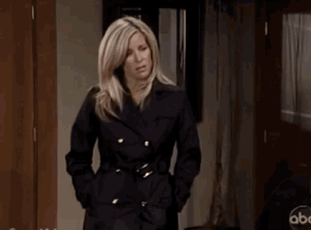 a woman in a black trench coat is standing in a hallway with her hands in her pockets .