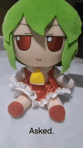 a stuffed doll with green hair and red eyes is sitting on a table with the words `` asked '' below it .