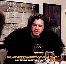 a man in a fur coat is sitting at a table with a glass of wine and talking to someone .