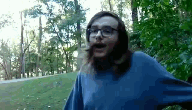 a man wearing glasses and a blue sweater is standing in the woods .