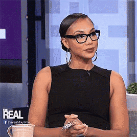 a woman wearing glasses and a black tank top is sitting in front of a sign that says real