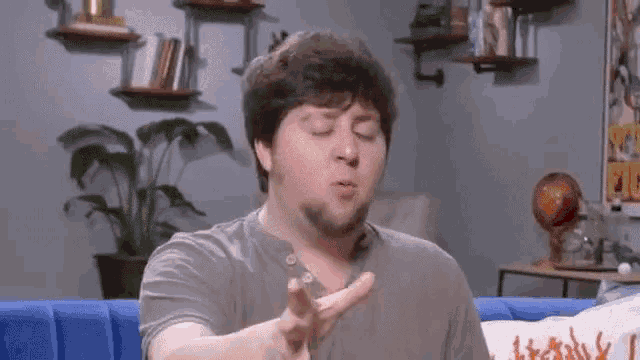 a man with a beard is sitting on a blue couch making a gesture with his hand .