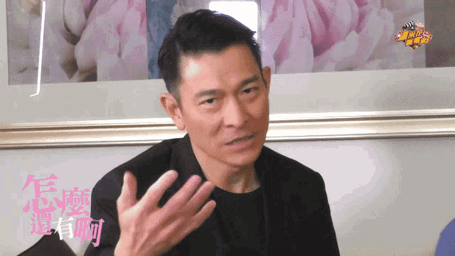 a man in a black suit talks to someone in front of a framed picture of pink flowers