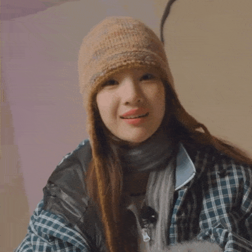 a woman wearing a plaid jacket and a knitted hat is smiling and wearing a scarf around her neck
