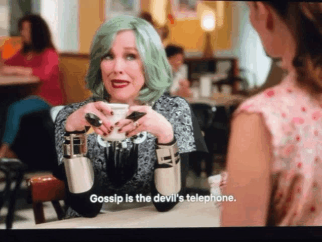 a woman with green hair is sitting at a table holding a cup of coffee and says gossip is the devil 's telephone