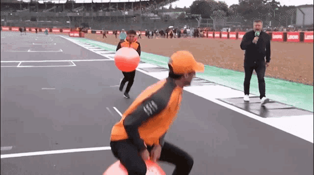 a man is riding a bouncy ball on a race track while another man holds an orange balloon .