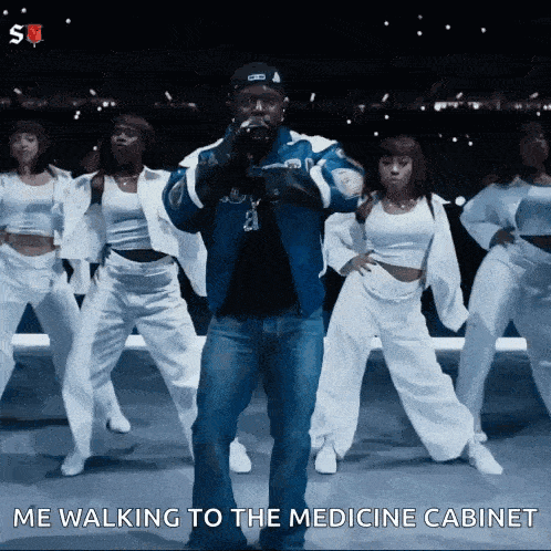 a man in a blue jacket is standing in front of a group of dancers with the caption " me walking to the medicine cabinet "