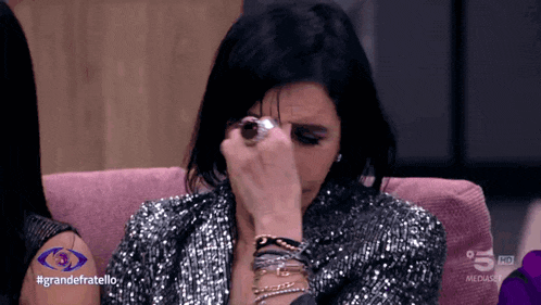 a woman in a sequined jacket wipes her eyes while sitting on a pink couch