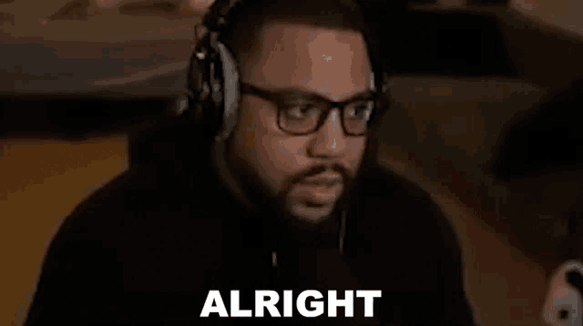 a man wearing headphones and glasses is sitting in front of a computer and saying `` alright '' .