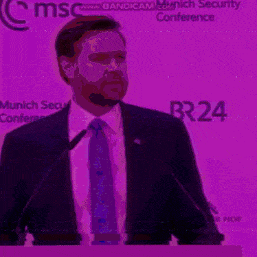 a man in a suit and tie stands at a podium in front of a wall that says br 24