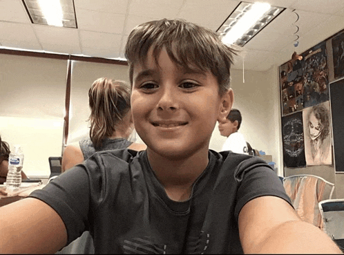 a young boy taking a selfie in a classroom with a bottle of aquafina water behind him