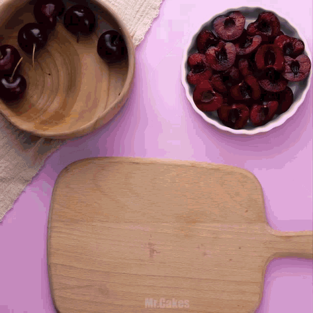 a wooden cutting board sits on a pink table next to a bowl of cherries and a bowl of raspberries