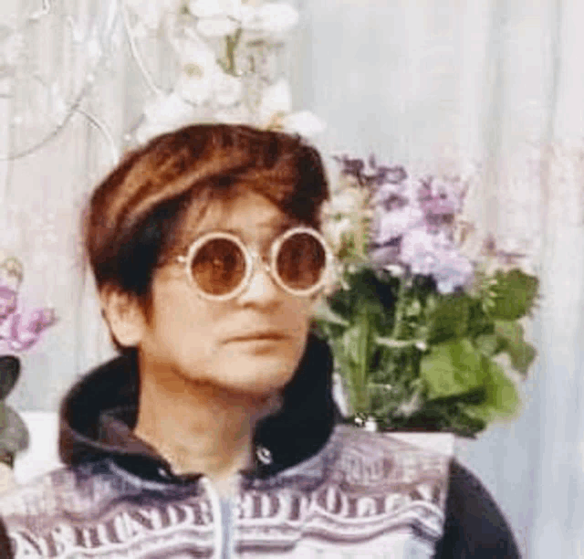 a woman wearing sunglasses and a sweater is standing in front of a vase of flowers .