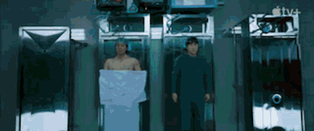 two men are standing next to each other in a room with a towel wrapped around their torsos .