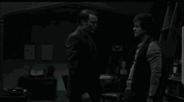 two men standing next to each other in a dark room with a clock on the wall
