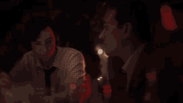 two men are sitting at a table in a dark room with red lights behind them