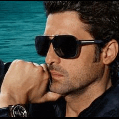 a man wearing sunglasses and a watch is sitting in front of the ocean .