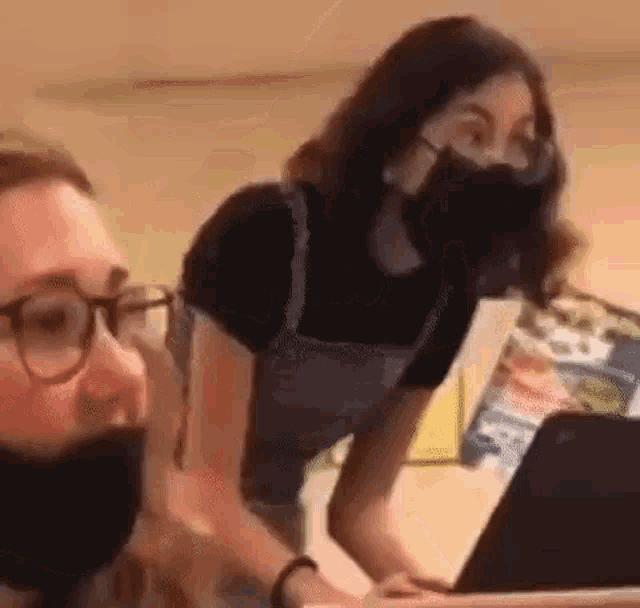 a woman wearing a mask is standing next to a man wearing glasses and a laptop .