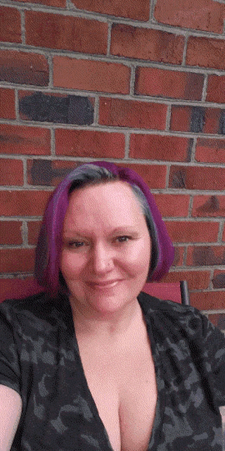 a woman with purple hair and gray eyebrows is smiling in front of a brick wall