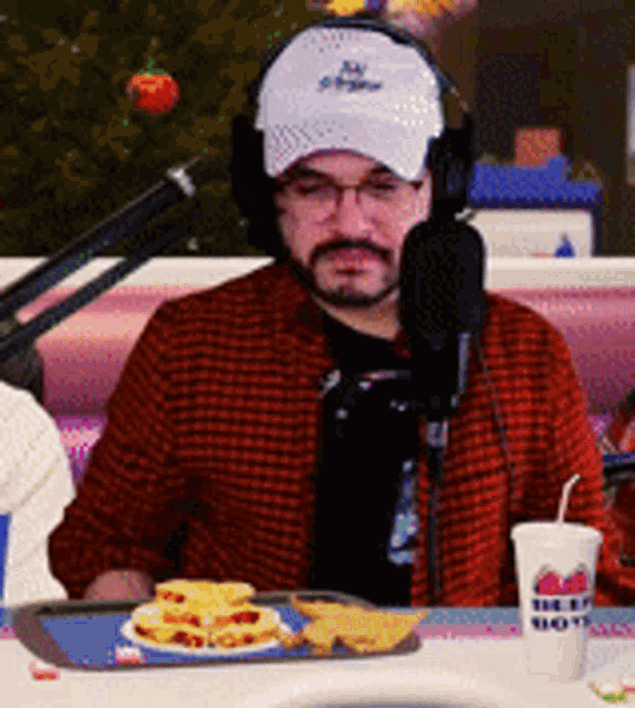 a man wearing a hat and headphones is sitting at a table with a microphone and a cup that says the big shot