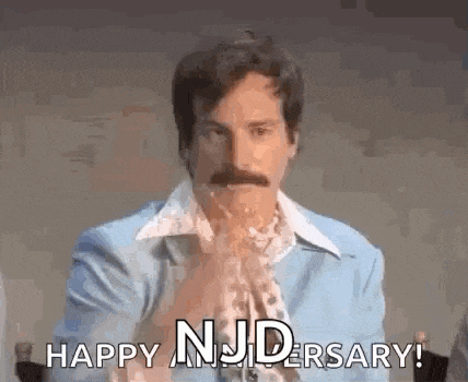 a man with a mustache is sitting at a table with his hands folded and says `` happy nj anniversary ! ''