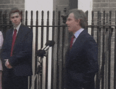 a man in a suit and tie stands in front of a fence