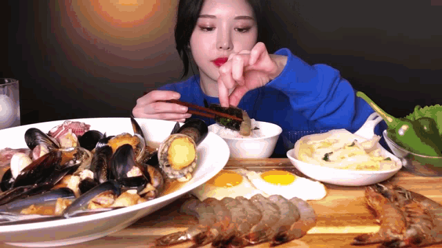a woman in a blue sweater is eating a plate of food with chopsticks