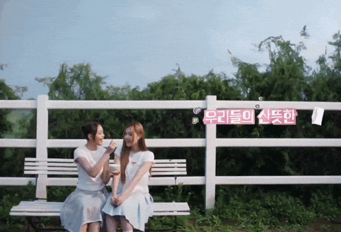 two women sit on a bench in front of a white fence with a sign that says ' 우리들 의 신붓한 '