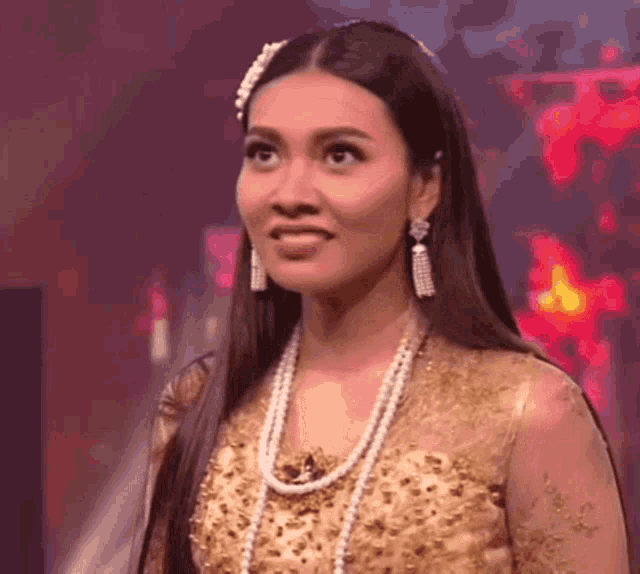 a woman with long hair is wearing a gold dress and pearls and earrings .