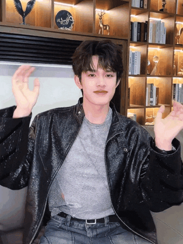 a young man wearing a black leather jacket and a grey shirt is sitting in front of a bookshelf