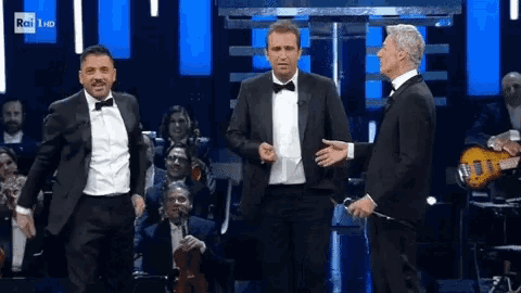 three men in tuxedos are standing on a stage in front of an orchestra with rai 1 hd written on the bottom of the screen