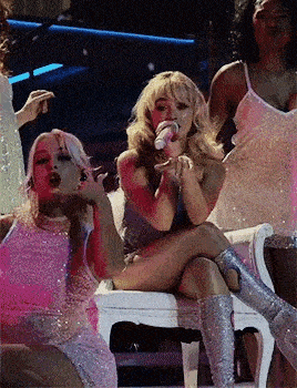 a woman is sitting on a chair singing into a microphone while a group of women are dancing around her .
