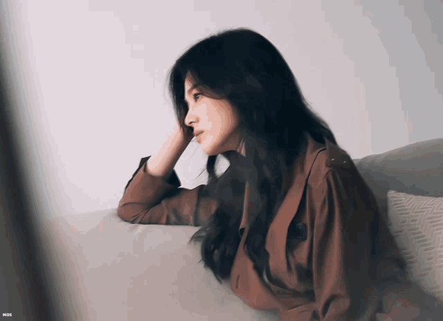 a woman in a brown jacket sits on a couch with her hand on her head