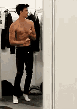 a shirtless man is standing in a dressing room looking at himself in a mirror .