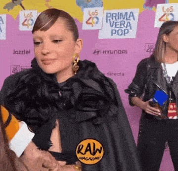 a woman in a black cape is standing in front of a pink wall that says prima vera pop