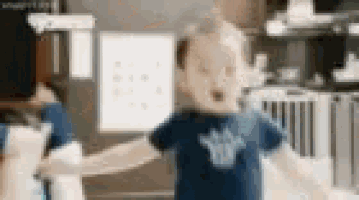 a baby in a blue shirt is standing in a kitchen holding a paper towel .