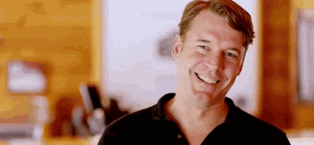 a man in a black shirt is smiling in front of a wooden wall