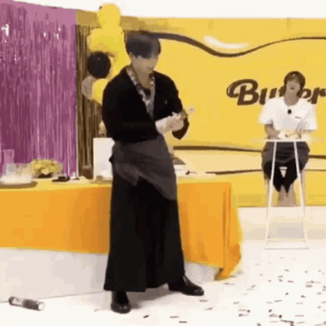 a man in a black dress is standing in front of a table holding a bottle .