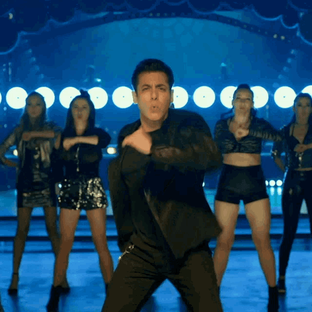 a man in a black jacket stands in front of a group of dancers