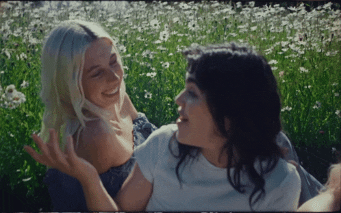 two women are sitting in a field of flowers and laughing