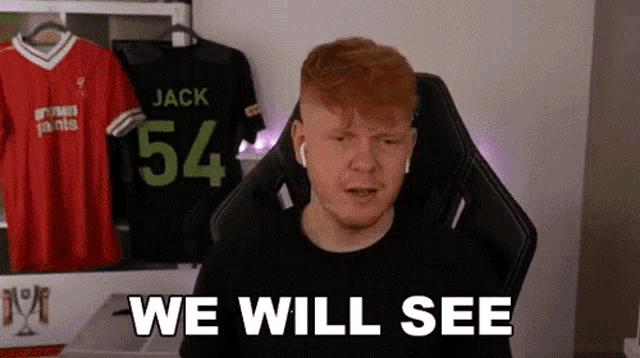 a man is sitting in a gaming chair and saying `` we will see '' in front of a wall with jerseys hanging on it .