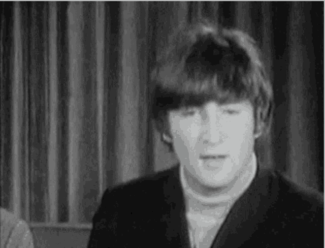 a black and white photo of john lennon making a funny face while sitting in front of a curtain .