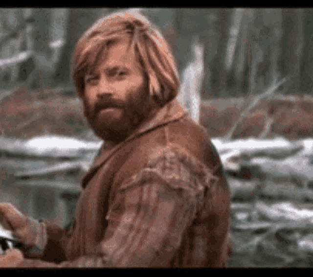 a man with a beard and long hair is standing in the woods holding a camera .