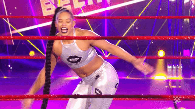 a woman in a silver top and white pants is standing in a wrestling ring with her arms outstretched .