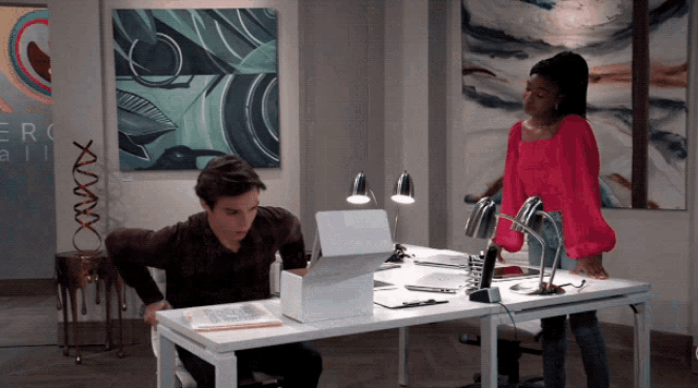 a woman in a pink shirt stands next to a man sitting at a desk with a laptop