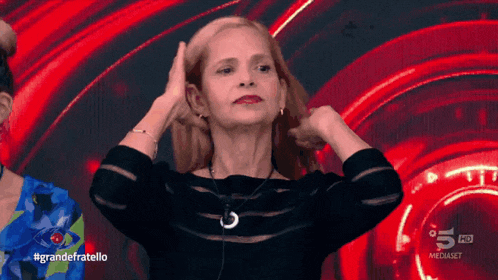 a woman adjusts her hair in front of a screen that says #grandefratello on it