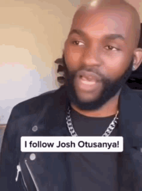 a man with a beard is wearing a black jacket and a chain around his neck and says i follow josh otusanya .